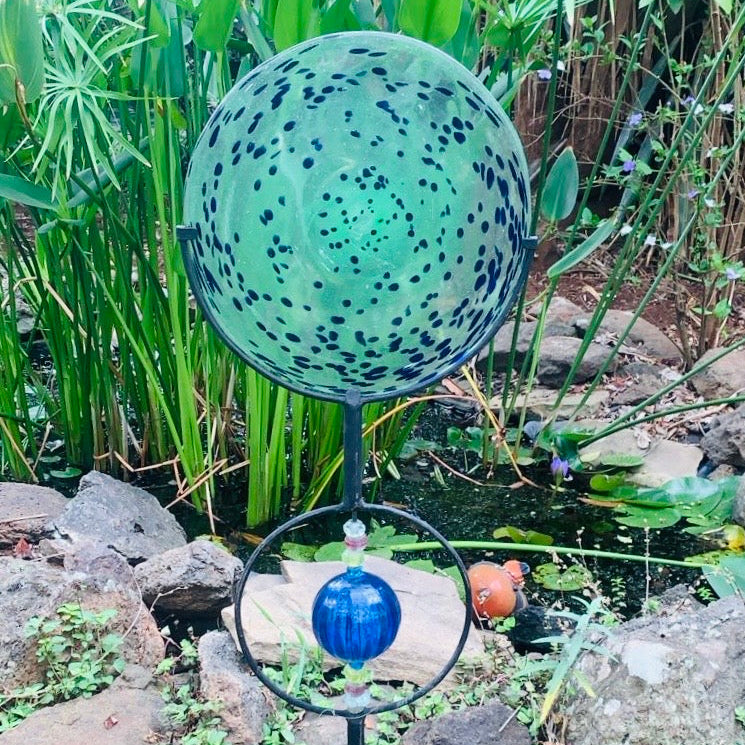 Decorative garden sphere with blue and green speckled design on a stand, surrounded by greenery and rocks.