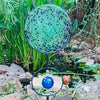 Decorative garden sphere with blue and green speckled design on a stand, surrounded by greenery and rocks.