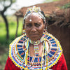 A close-up portrait of a masai woman wearing traditional African beaded jewelry, including a large circular beaded collar (omwe), layered necklaces, and a headpiece, with a red patterned shawl over a white top, set against a blurred outdoor background.