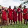 A group of Maasai men standing outdoors in front of a traditional earth hut, wearing bright red shawls and body paint, holding wooden sticks, with grass and trees in the background.