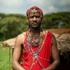 A man wearing traditional red clothing and elaborate multicolored beaded necklaces, bracelets, and a red headwrap stands outdoors in front of a thatched structure under a cloudy blue sky.