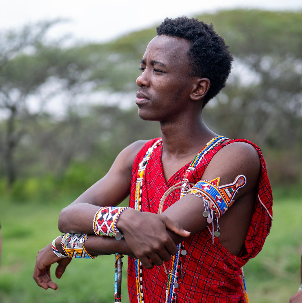A man wearing traditional Maasai attire, including a red patterned wrap, beaded necklaces, bracelets, and a black waist sash, standing outdoors in a grassy field.
