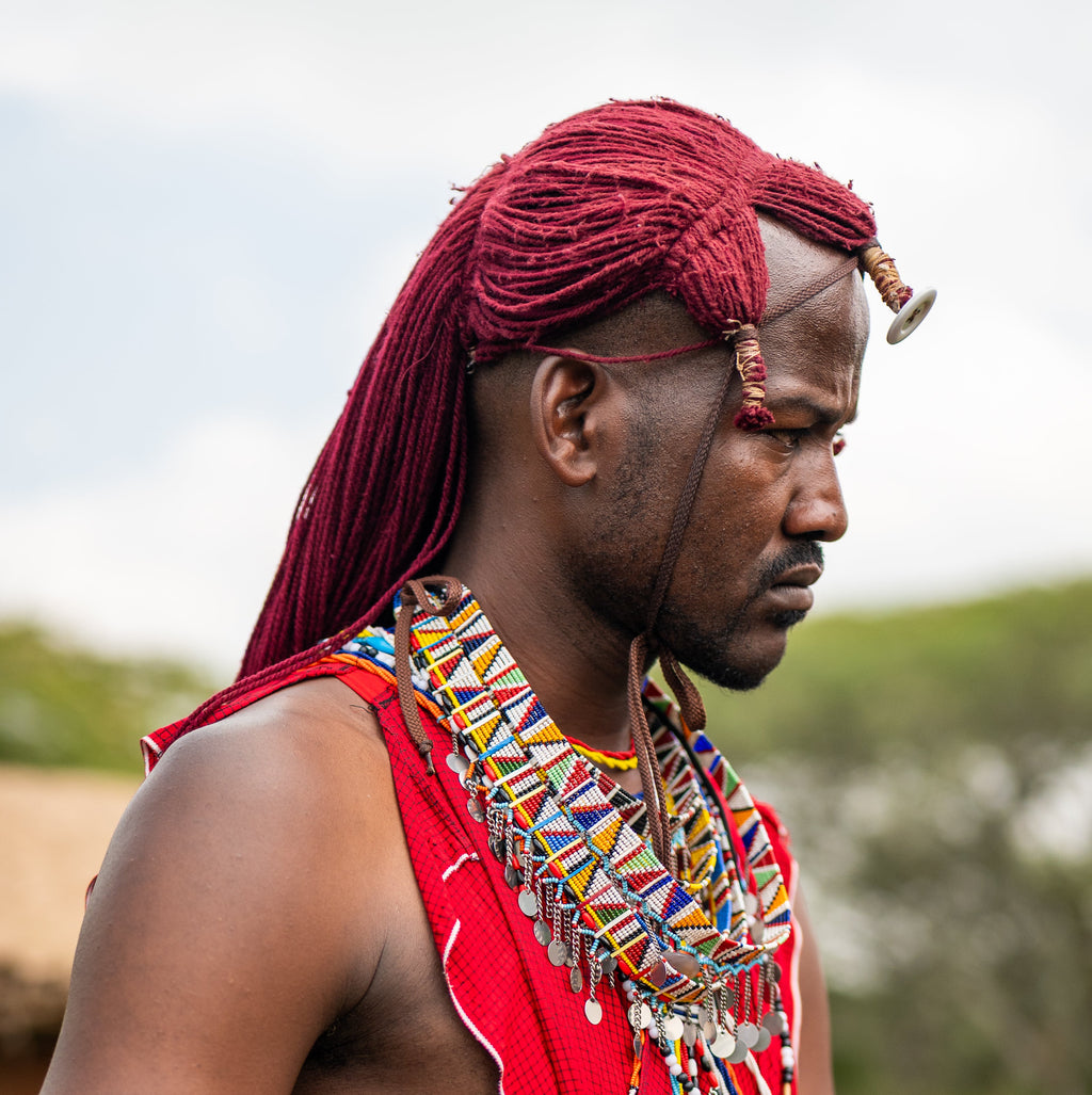 A man wearing traditional red Maasai-style clothing, layered beaded necklaces, bracelets, and a red headwrap, standing outdoors with blurred hills and a hut in the background.