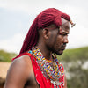 A man wearing traditional red Maasai-style clothing, layered beaded necklaces, bracelets, and a red headwrap, standing outdoors with blurred hills and a hut in the background.