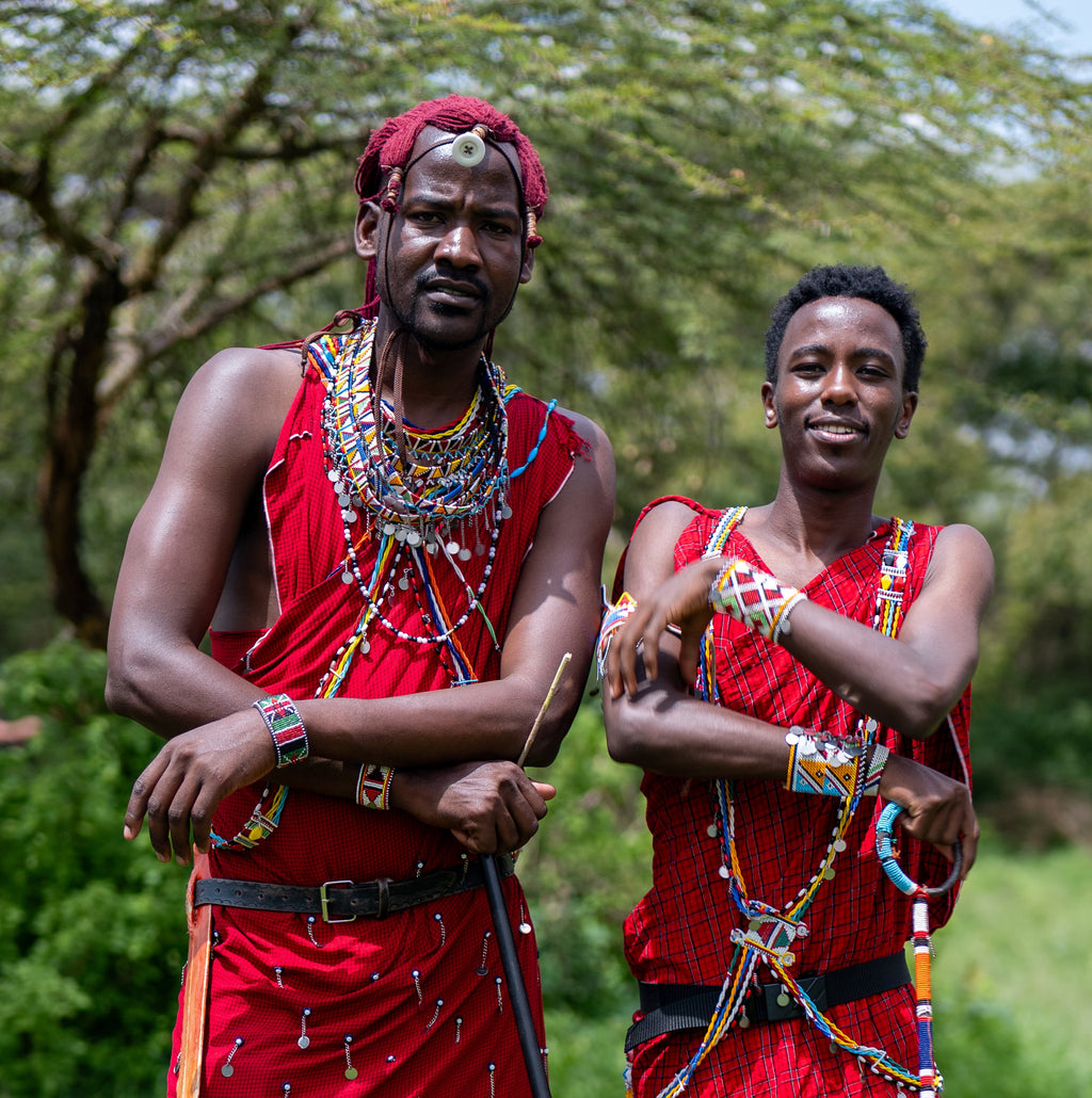 Two people standing outdoors in grassy terrain wearing bright red traditional masai garments and layered beaded necklaces, with trees in the background.