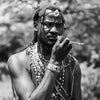 A black and white portrait of a man outdoors wearing traditional masai attire, including a beaded waist sash, necklaces, and a circular forehead ornament, while holding a small object near his mouth.